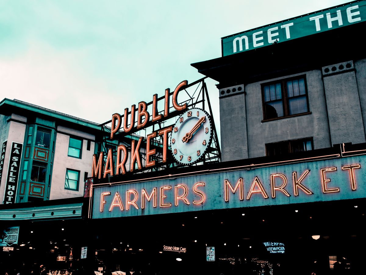 The image shows a neon sign for a "PUBLIC MARKET" and "FARMERS MARKET" with buildings in the background, possibly in a busy urban area.