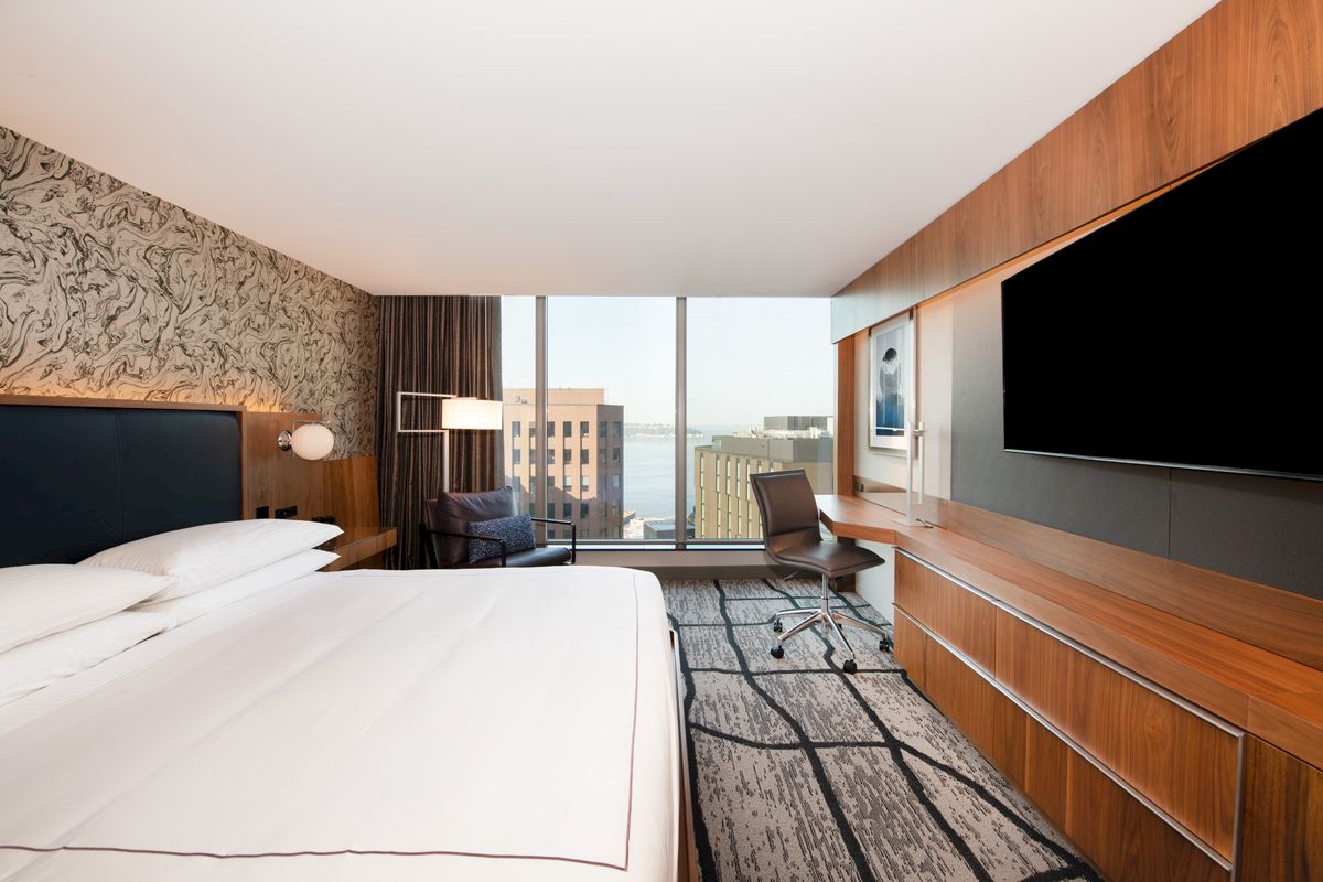 A modern hotel room with a large bed, flat-screen TV, desk, chair, and a view of a cityscape through tall windows in the background.