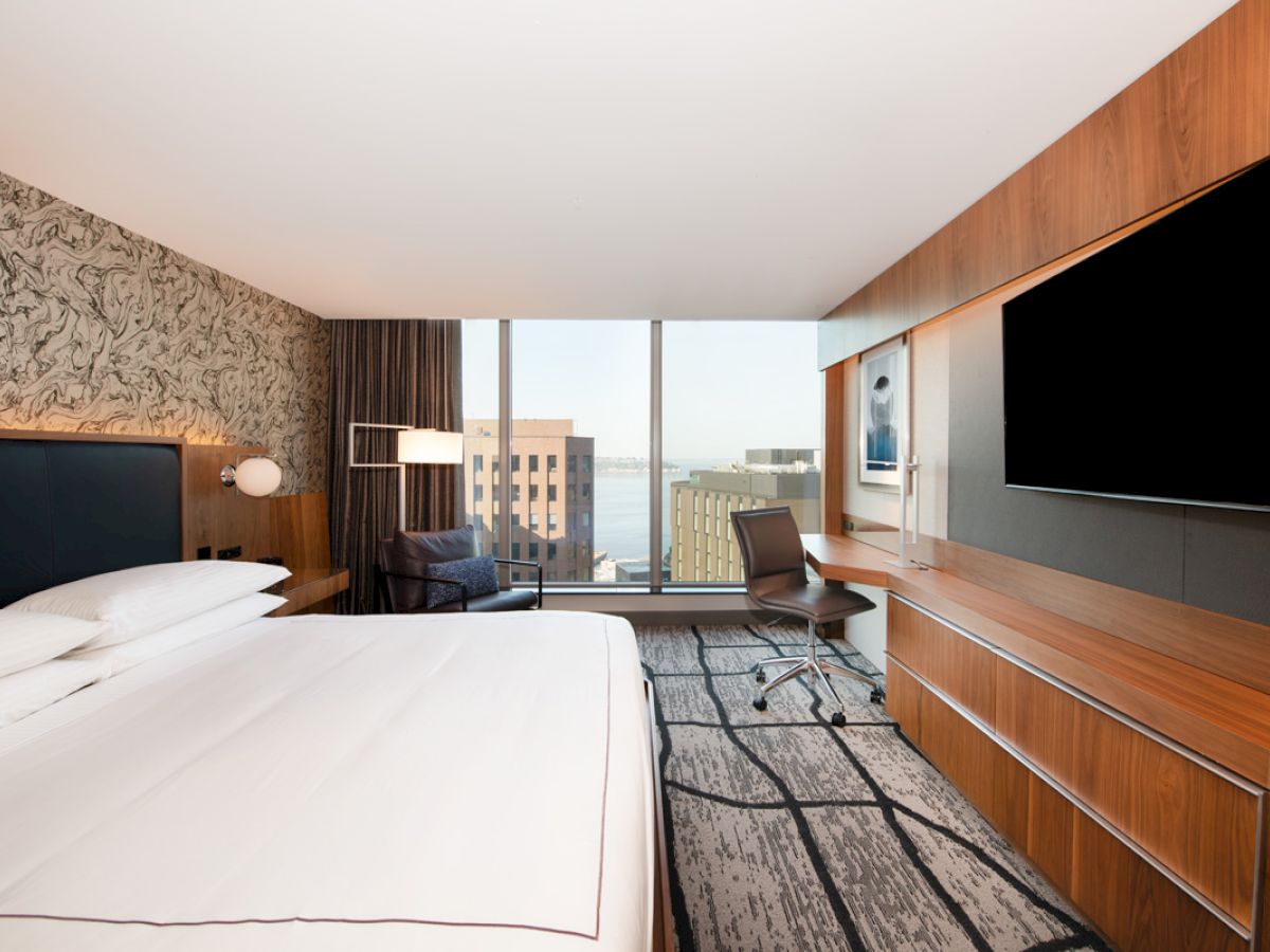 A modern hotel room with a large bed, flat-screen TV, desk, chair, and a view of a cityscape through tall windows in the background.