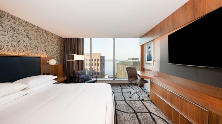 A modern hotel room with a large bed, flat-screen TV, desk, chair, and a view of a cityscape through tall windows in the background.