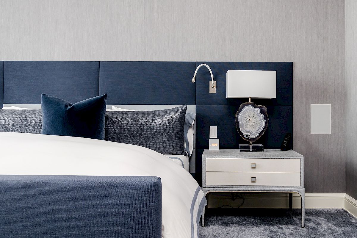 A modern bedroom features a blue upholstered bed, two dark blue pillows, a nightstand with a lamp, decorative items, and a gray carpet.