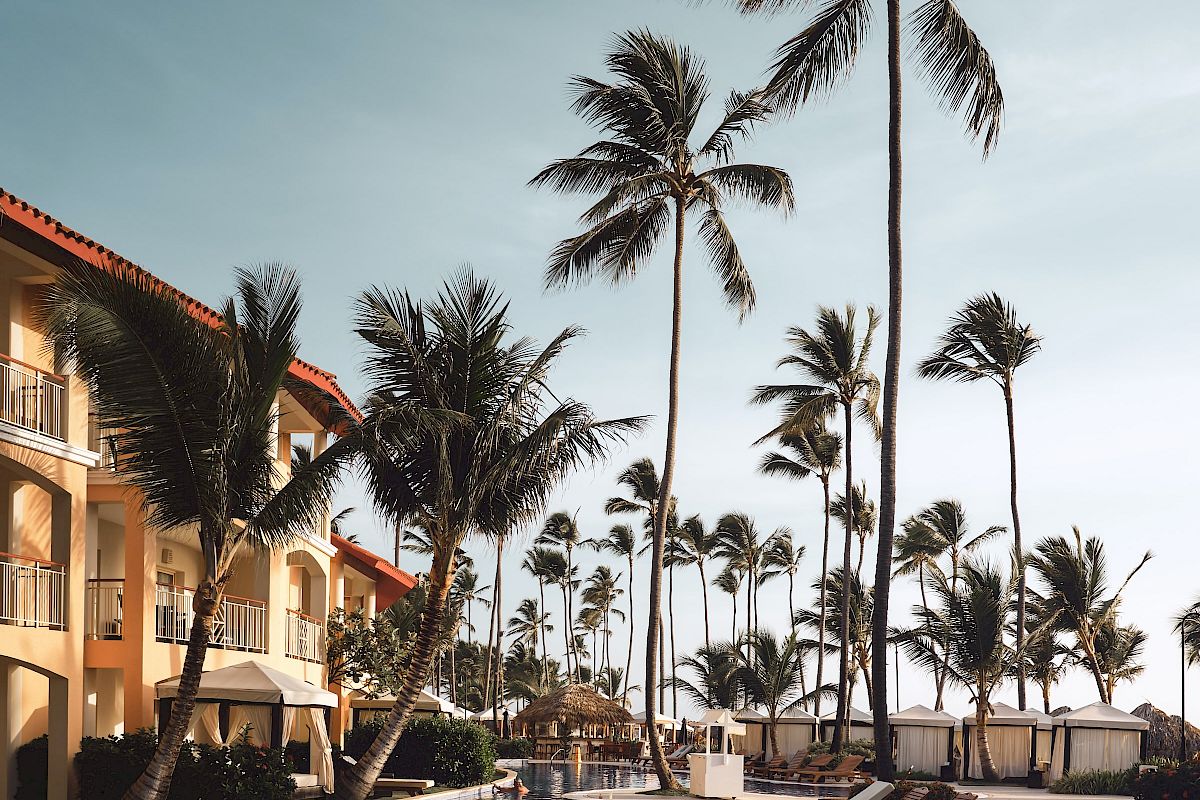 This image shows a poolside scene with lounge chairs, tall palm trees, and a building with balconies under a clear blue sky.