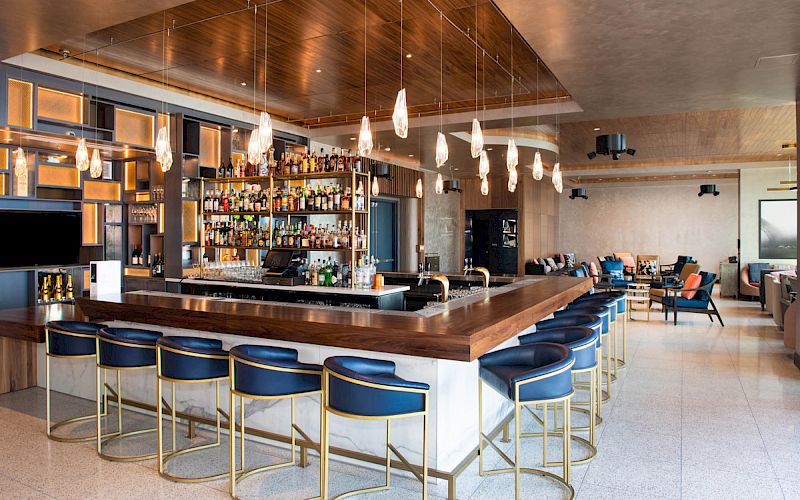 The image shows a modern bar with stylish high stools, pendant lighting, a fully stocked bar, and a cozy seating area in the background, ending the sentence.
