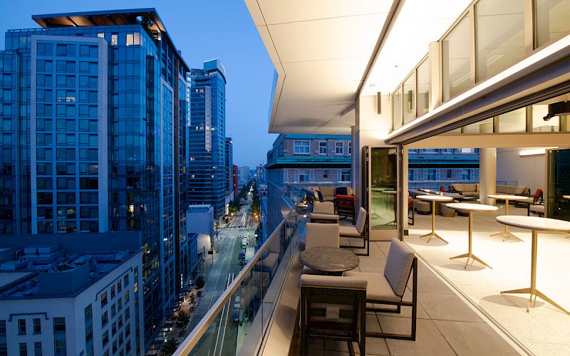 The image shows a modern cityscape at dusk with tall buildings, a street below, and a furnished open balcony with seating and tables.