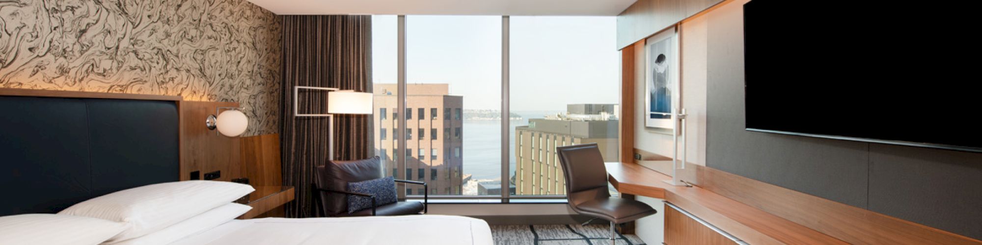 A modern hotel room with a large bed, wall-mounted TV, desk and chair, and a window with a view of buildings and the sea in the distance.