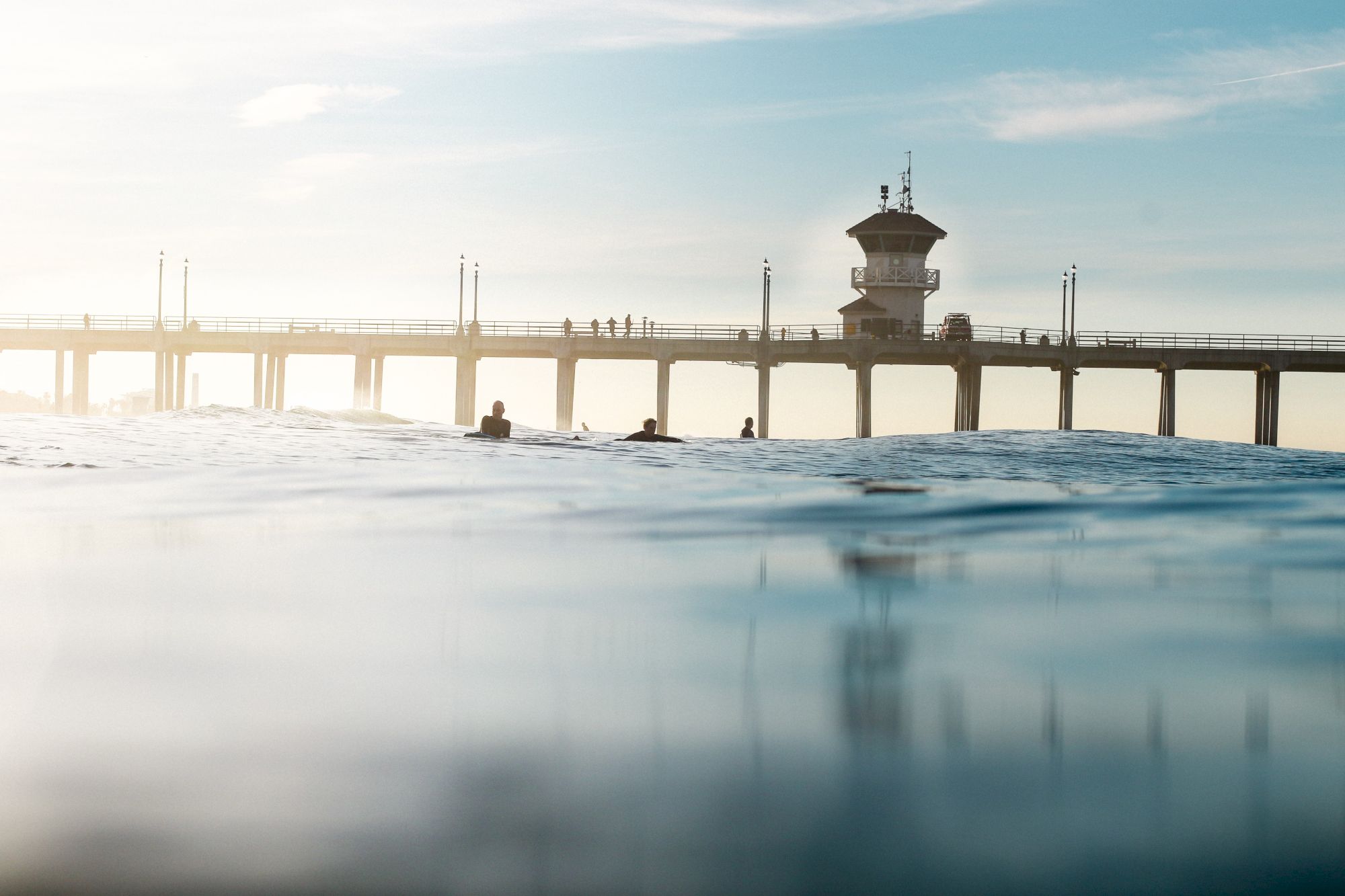 The image shows a pier extending over a calm ocean, with people standing and walking on the pier, and a lighthouse-like structure in the background.